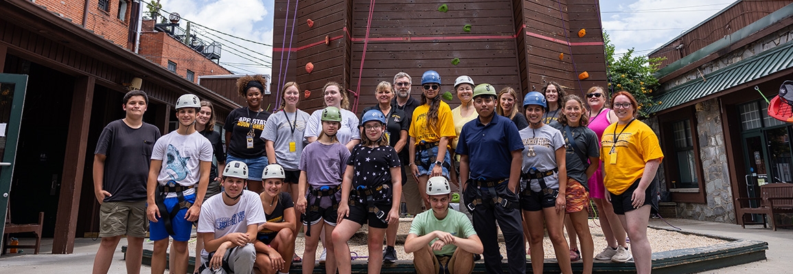 Group of kids, students and faculty stand in front of a large climbing tower