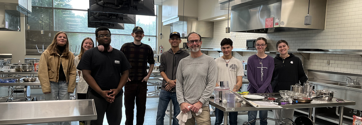 Group of smiling individuals stand in a line inside a large, stainless steel cooking space 