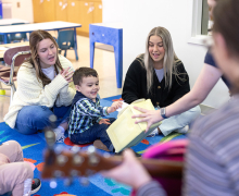 App State speech-language pathology graduate students Peyton Arrowood, of Rutherfordton, left, and Ellie Bowen, of Hendersonville, work with a young child at the university’s Thomas O. Eller Preschool Language Classroom, guiding the child through a speech therapy exercise. Photo by Chase Reynolds Two younger women sit on brightly colored carpeted squares behind a toddler with dark hair while all three are clapping
