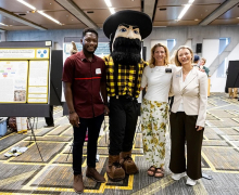 Three people stand with a mascot in the middle of them inside a classroom setting 
