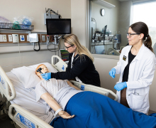 Woman wearing scrubs using a medical instrument over a simulation manikin's face while another female practitioner in a white lab coat observes. 