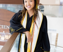 Graduate wears cap and gown and stands in lobby that says Health Sciences 