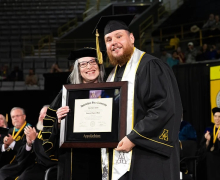 A woman and man wearing black commencement regalia stand together with the man holding a framed diploma 