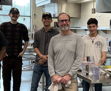 Group of four individuals stand inside a stainless steel, large commercial kitchen 