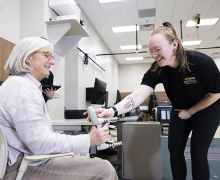 Woman squeezes a grip measurement device while a younger woman in a black App State polo holds out her hand to help