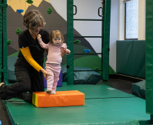 Young child steps on an orange colored mat with the help of a woman who is standing behind her and holding her legs for support. 
