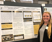 Young woman with long, blonde hair stands beside a research poster with different text boxes and writing on it 