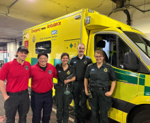 Group of five people wearing official looking logoed shirts stand side by side in front of a yellow ambulance 