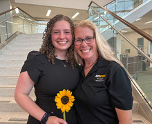 Young woman with curly dark hair holds a sunflower while standing next to another woman with blonde hair; both are smiling widely 