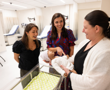 Two young women look at a human baby simulator being held by a woman in a white coat. 
