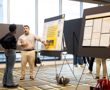 Person points to a poster on an easel while talking to someone else in a large room with other posters. 