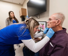 Woman in medical scrubs put her hands near the throat of an older adult man who is sitting in a clinic room