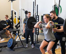 Four people are inside a gym space working with free weights; a young woman with a ponytail is lifting a large weighted bar while a man stands behind her spotting her. 