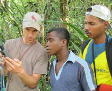 Man holds an instrument in a forested area with two other men looking over his shoulder 