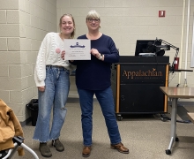 Two women are standing next to each other and are smiling; one is holding a certificate. 
