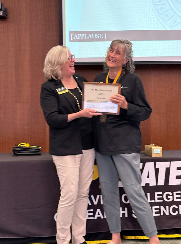 Two women stand in a room and look at each other; one is holding a framed certificate