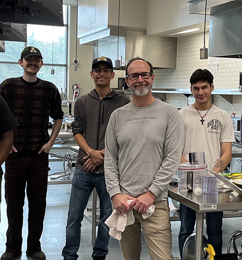 Group of four individuals stand inside a stainless steel, large commercial kitchen 