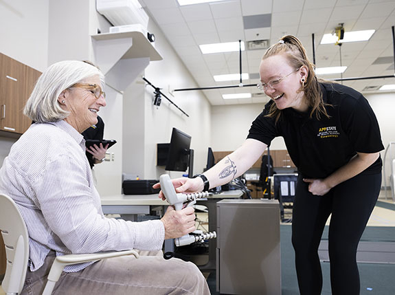 Woman squeezes a grip measurement device while a younger woman in a black App State polo holds out her hand to help