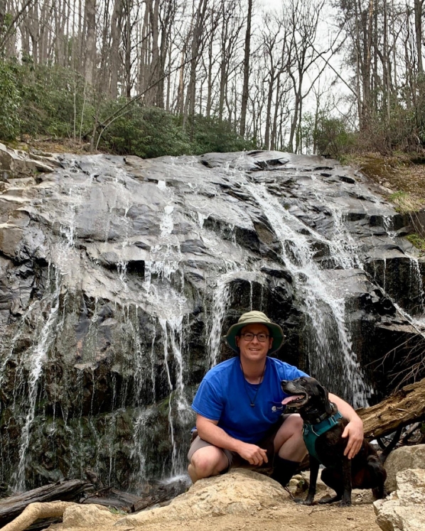 Photo of man standing in front of a rock waterfall with a black dog 