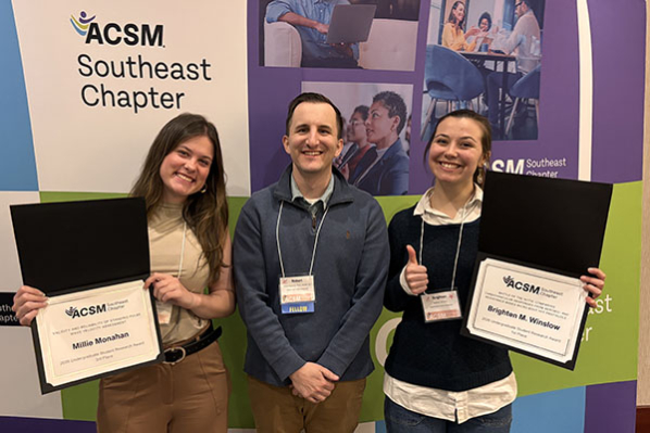 Two women hold certificates with a man standing between them and a banner behind them that reads SEACSM conference