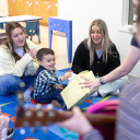 Two younger women sit on brightly colored carpeted squares behind a toddler with dark hair while all three are clapping