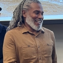 Smiling man with salt and pepper dreadlocks wears a brown shirt 
