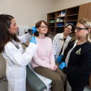 Women in white coat looks inside a woman's ear while two other clinical people look on. 