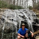 Photo of man standing in front of a rock waterfall with a black dog 