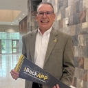 Man wearing a blazer smiles as he stands inside the lobby area of a building 