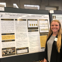 Young woman with long, blonde hair stands beside a research poster with different text boxes and writing on it 