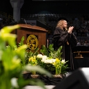 Woman wearing graduation robes smiles and claps from stage 