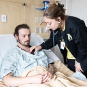 Woman uses a stethoscope to listen to a person wearing a gown in a hospital bed's heartbeat
