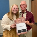 Student is standing beside a retired professor holding a sign that says Classroom 