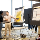 Person points to a poster on an easel while talking to someone else in a large room with other posters. 