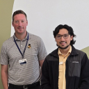 Two people stand in front of a green wall with a white graphic; one is wearing a lanyard and a name badge