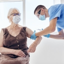 Person in scrubs leans over to give a shot to an older woman sitting in a chair 