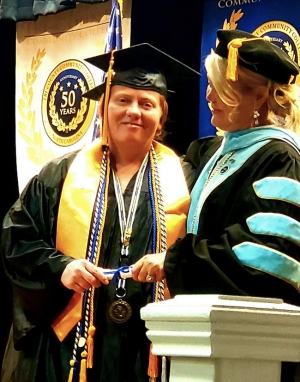 Woman wearing a graduation gown and mortar board while being presented with a degree