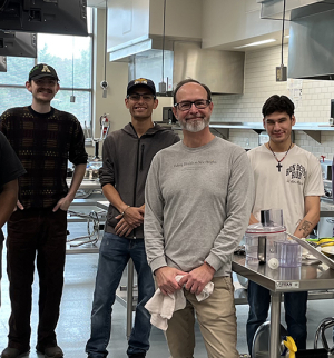 Group of four individuals stand inside a stainless steel, large commercial kitchen 