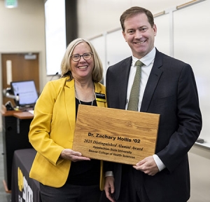 Two individuals stand together with one holding a wooden plaque