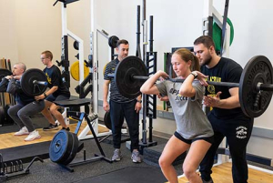 Four people are inside a gym space working with free weights; a young woman with a ponytail is lifting a large weighted bar while a man stands behind her spotting her. 