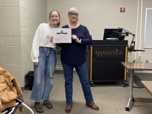 Two women are standing next to each other and are smiling; one is holding a certificate. 