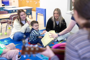 Two younger women sit on brightly colored carpeted squares behind a toddler with dark hair while all three are clapping