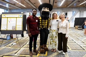 Three people stand with a mascot in the middle of them inside a classroom setting 