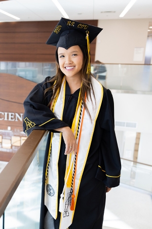 Graduate wears cap and gown and stands in lobby that says Health Sciences 