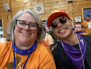 Woman in an orange shirt and bandana leans toward another person who is wearing sunglasses and a cap in an indoor rustic setting 
