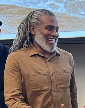 Smiling man with salt and pepper dreadlocks wears a brown shirt 