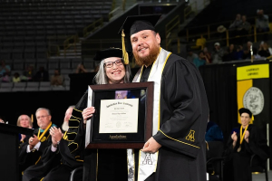 A woman and man wearing black commencement regalia stand together with the man holding a framed diploma 