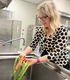 Woman wearing a polka dotted sweater washes carrots in an industrial sink.