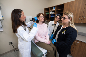 Women in white coat looks inside a woman's ear while two other clinical people look on. 
