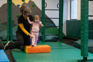 Young child steps on an orange colored mat with the help of a woman who is standing behind her and holding her legs for support. 